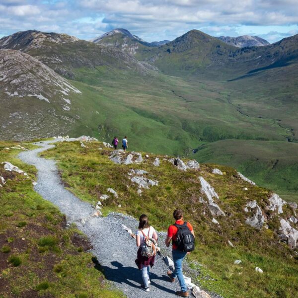 Several hikers walk along a gravel trail through a green mountainous landscape under a partly cloudy sky.