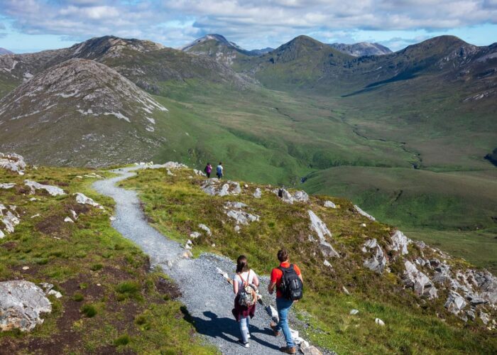 Several hikers walk along a gravel trail through a green mountainous landscape under a partly cloudy sky.