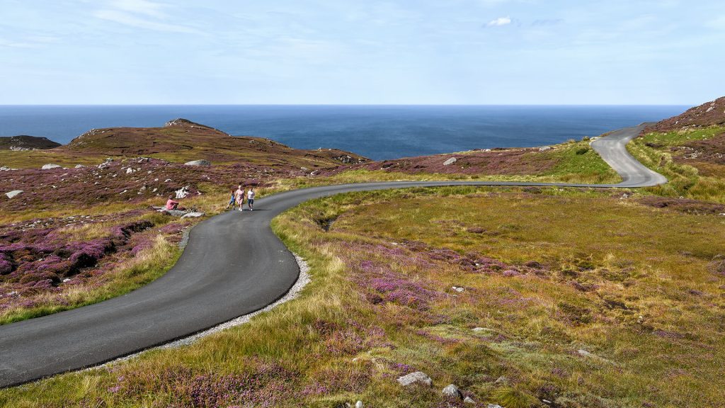 A winding road on a hilly landscape leads towards the sea. People are walking along the road under a clear sky.