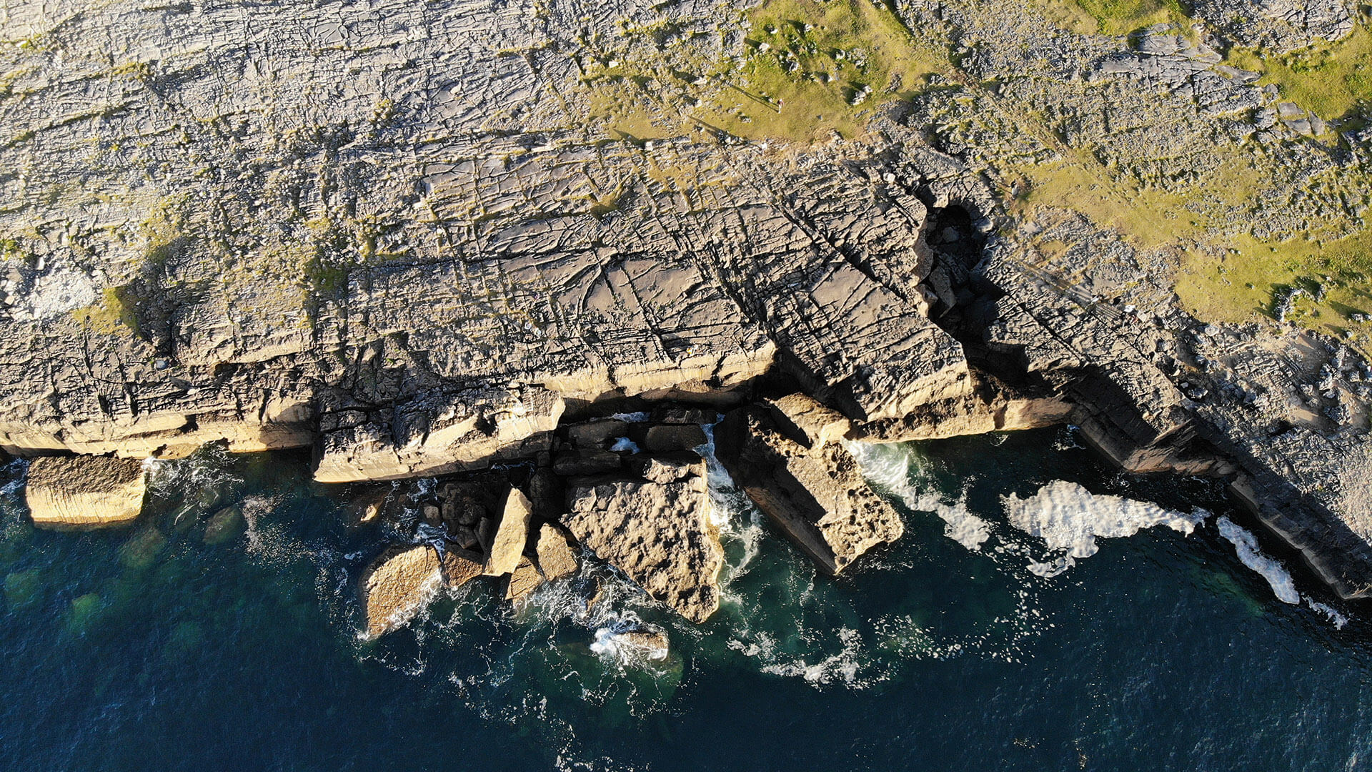 Aerial view of rocky cliffs meeting clear, dark blue water. The cliffs have deep cracks and jagged edges with sparse vegetation growing on top.