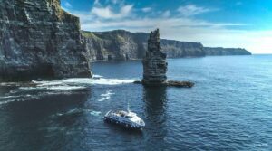 A tour boat sails near the coastal cliffs and a tall sea stack under a clear blue sky on a Cliffs Cruise from Galway to the Aran Islands.
