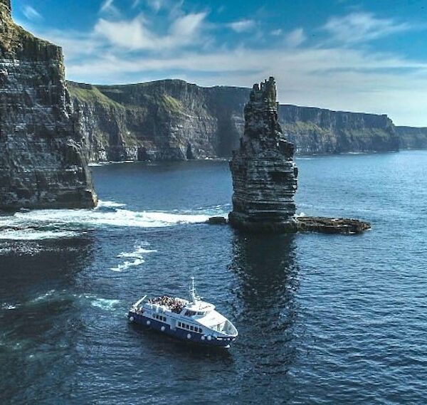 A tour boat sails near the coastal cliffs and a tall sea stack under a clear blue sky on a Cliffs Cruise from Galway to the Aran Islands.