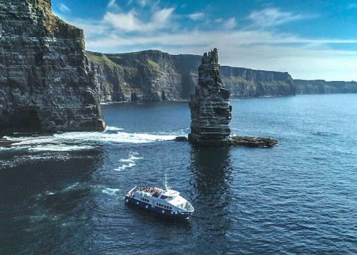 A tour boat sails near the coastal cliffs and a tall sea stack under a clear blue sky on a Cliffs Cruise from Galway to the Aran Islands.