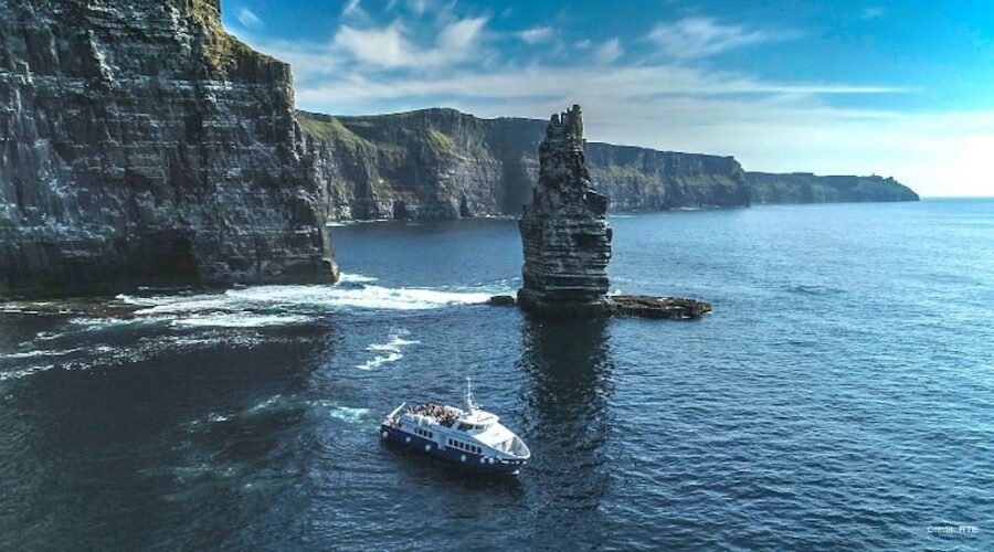 A tour boat sails near the coastal cliffs and a tall sea stack under a clear blue sky on a Cliffs Cruise from Galway to the Aran Islands.