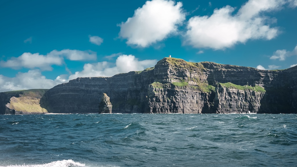 Steep rocky cliffs with patches of grass rise above the ocean near Galway, under a partly cloudy blue sky—an unforgettable view for anyone on a Cliffs Cruise to the Aran Islands.