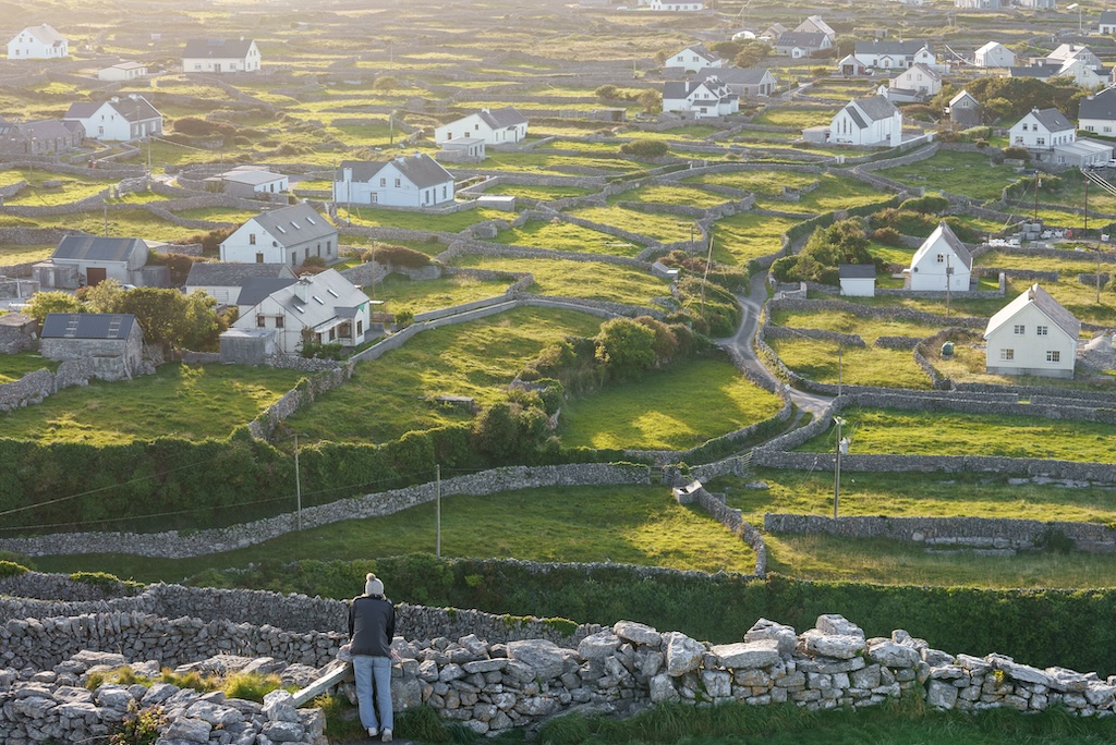 A person stands by a stone wall overlooking a rural Galway landscape with green fields, stone fences, and scattered white houses under soft sunlight.