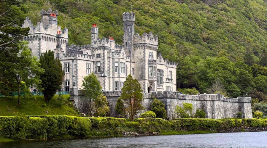 A large stone castle with towers, known as Kylemore Abbey, sits beside a lake, surrounded by dense green trees and hills along the scenic Galway to Connemara route.