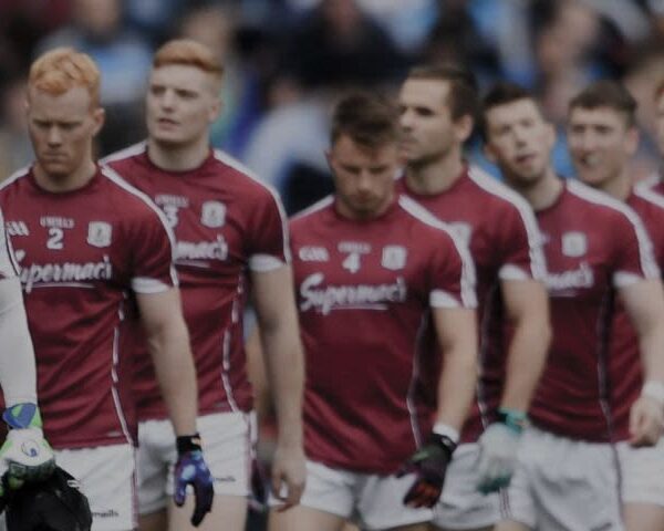 A group of male athletes in maroon jerseys stand in a line on a sports field, preparing for a game as the GAA Supporters Bus arrives in the background.
