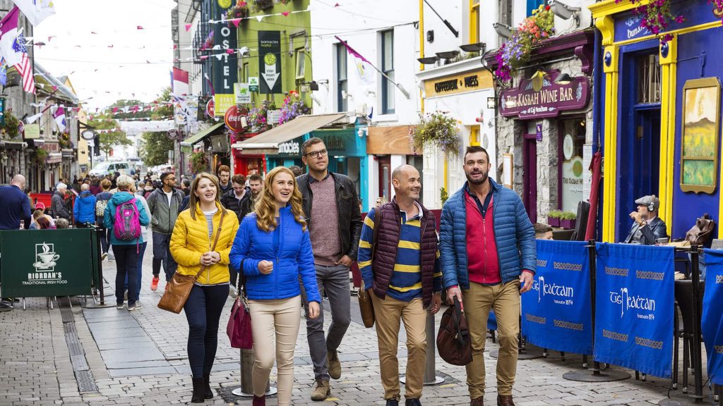 A group of people walk down a lively street lined with colorful buildings, shops, and cafes. The street is bustling with other pedestrians and decorated with hanging flowers and flags.