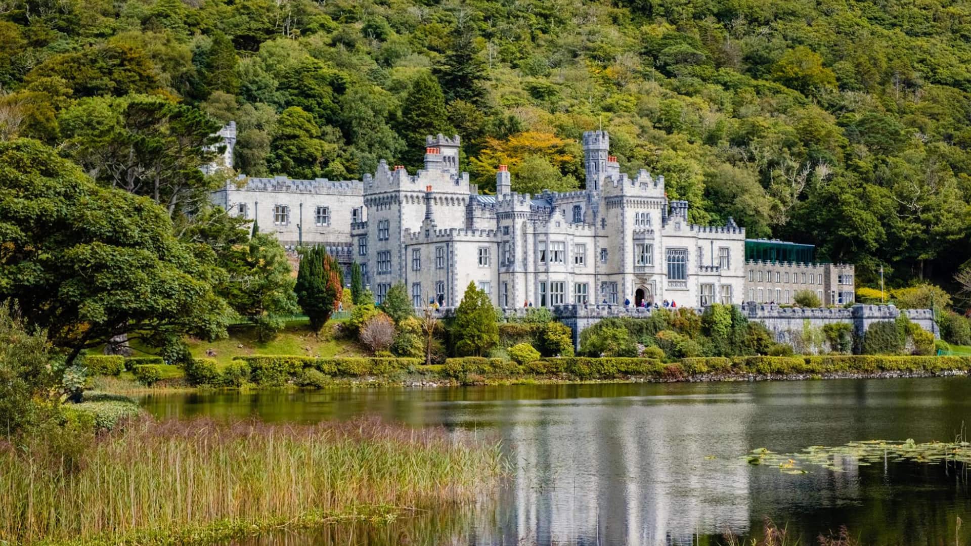 A large, historic stone castle with multiple towers and turrets is situated by a lake, surrounded by lush greenery.
