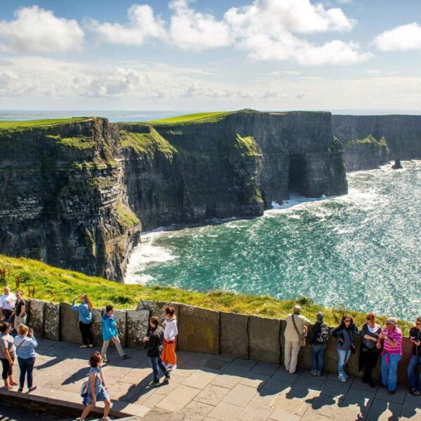 People stand on a viewing platform overlooking the Cliffs of Moher, featuring rugged cliffs and the Atlantic Ocean under a partly cloudy sky.