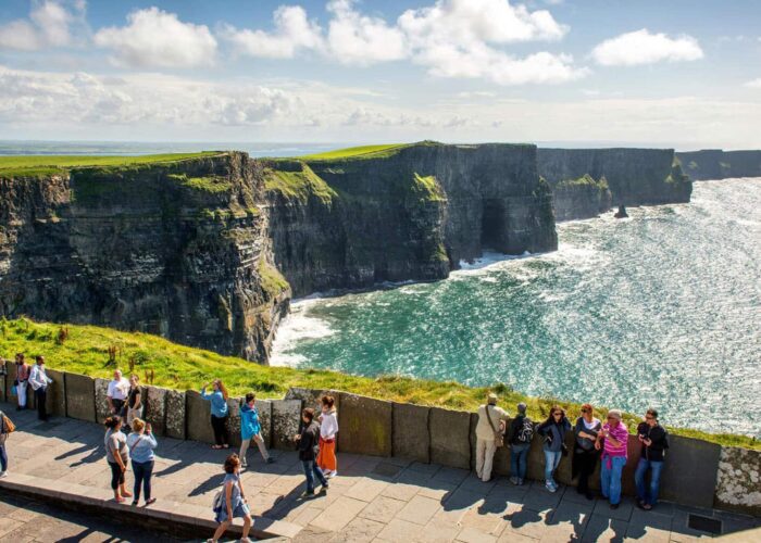 People stand on a viewing platform overlooking the Cliffs of Moher, featuring rugged cliffs and the Atlantic Ocean under a partly cloudy sky.