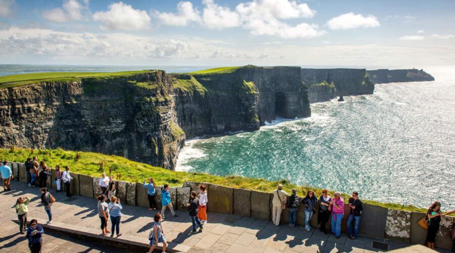 People stand on a viewing platform overlooking the Cliffs of Moher, featuring rugged cliffs and the Atlantic Ocean under a partly cloudy sky.