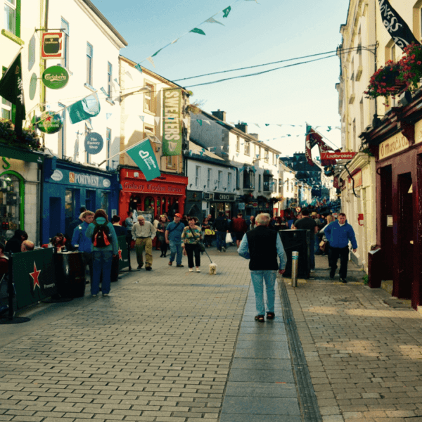 A bustling, narrow pedestrian street lined with colorful shops and restaurants, with people walking and sitting outdoors on a sunny day.