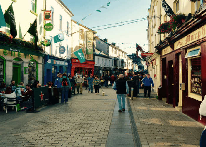 A bustling, narrow pedestrian street lined with colorful shops and restaurants, with people walking and sitting outdoors on a sunny day.