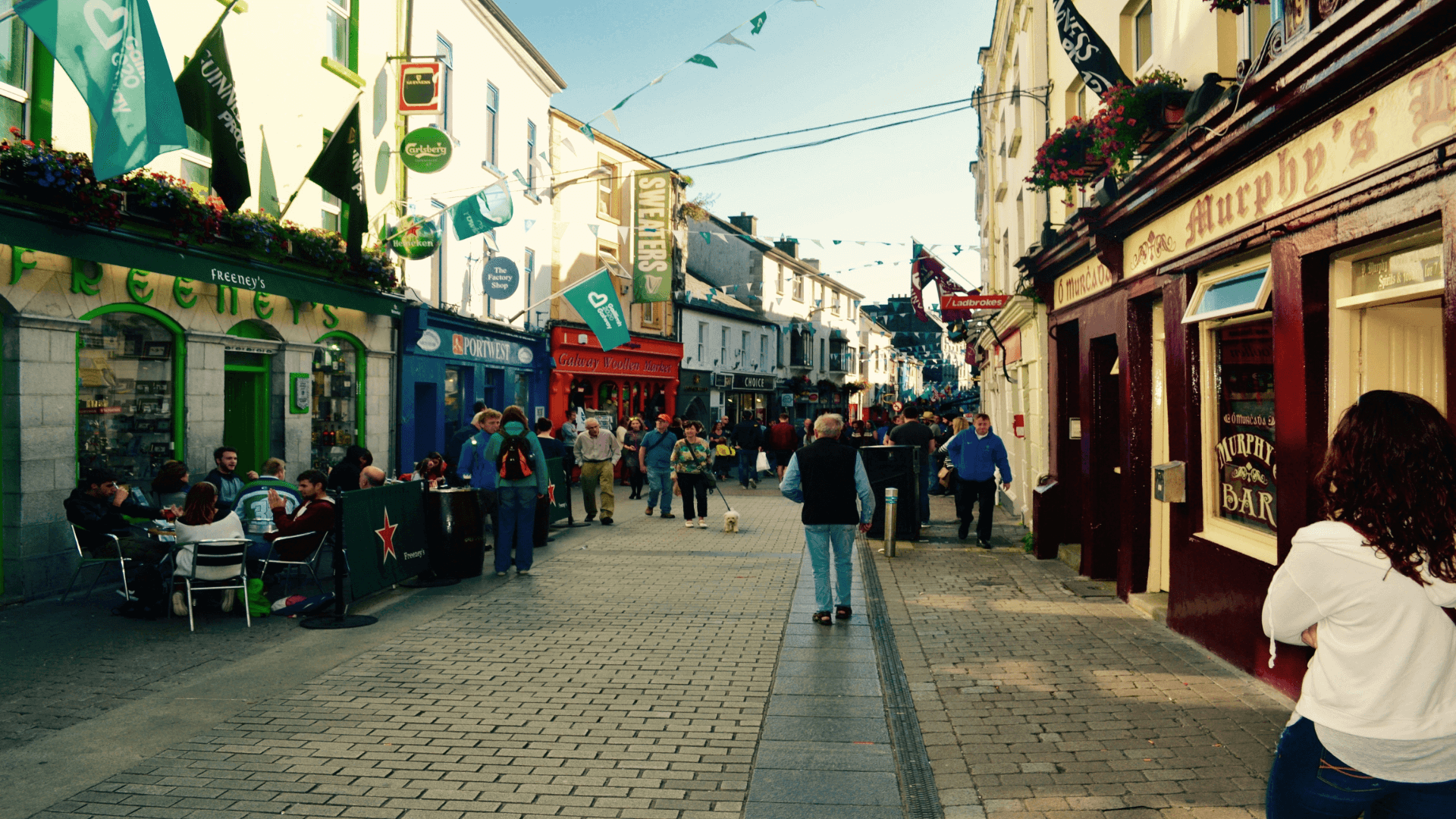 A bustling, narrow pedestrian street lined with colorful shops and restaurants, with people walking and sitting outdoors on a sunny day.