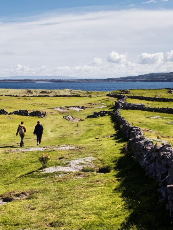Two people walk along a grassy path bordered by stone walls, with the sea and distant hills visible under a partly cloudy sky.