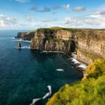 Steep cliffs covered in green grass overlook the blue ocean under a partly cloudy sky. On a 12 Day Ireland journey, watch waves crash gently against the base of these breathtaking cliffs.