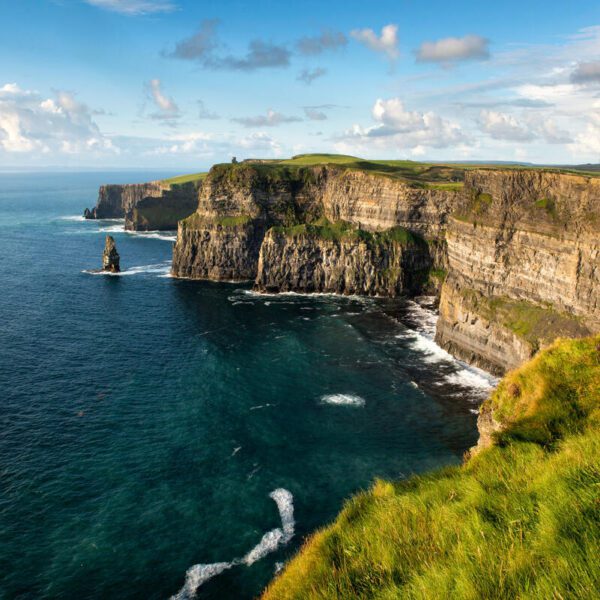 Steep cliffs covered in green grass overlook the blue ocean under a partly cloudy sky. On a 12 Day Ireland journey, watch waves crash gently against the base of these breathtaking cliffs.
