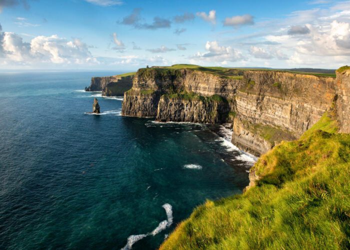 Steep cliffs covered in green grass overlook the blue ocean under a partly cloudy sky. On a 12 Day Ireland journey, watch waves crash gently against the base of these breathtaking cliffs.