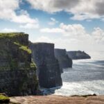 Steep sea cliffs rise above the ocean under a partly cloudy sky; people are visible walking along the grassy edge, experiencing the dramatic beauty featured on Ireland West tours.