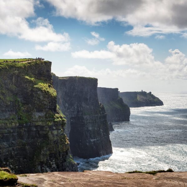 Steep sea cliffs rise above the ocean under a partly cloudy sky; people are visible walking along the grassy edge, experiencing the dramatic beauty featured on Ireland West tours.