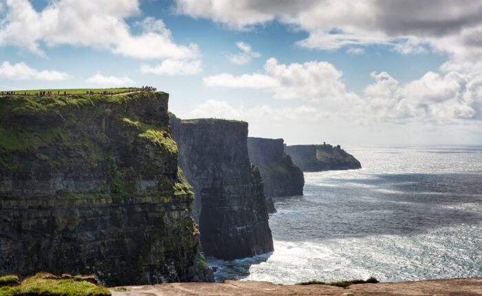 Steep sea cliffs rise above the ocean under a partly cloudy sky; people are visible walking along the grassy edge, experiencing the dramatic beauty featured on Ireland West tours.