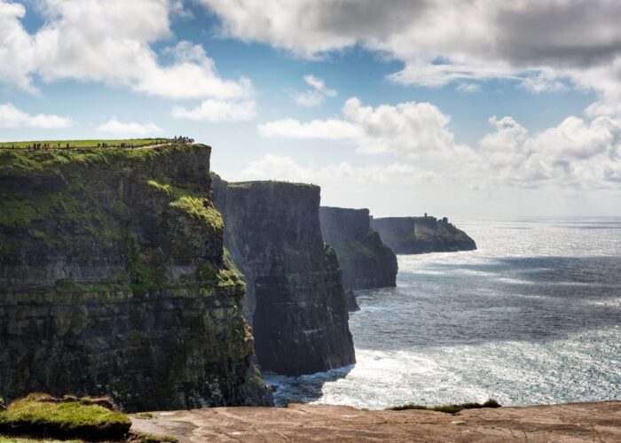 Steep sea cliffs rise above the ocean under a partly cloudy sky; people are visible walking along the grassy edge, experiencing the dramatic beauty featured on Ireland West tours.