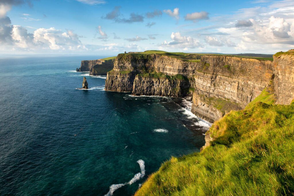 Cliffs rise steeply above the Atlantic Ocean, with green grass on top and waves crashing below under a partly cloudy sky.