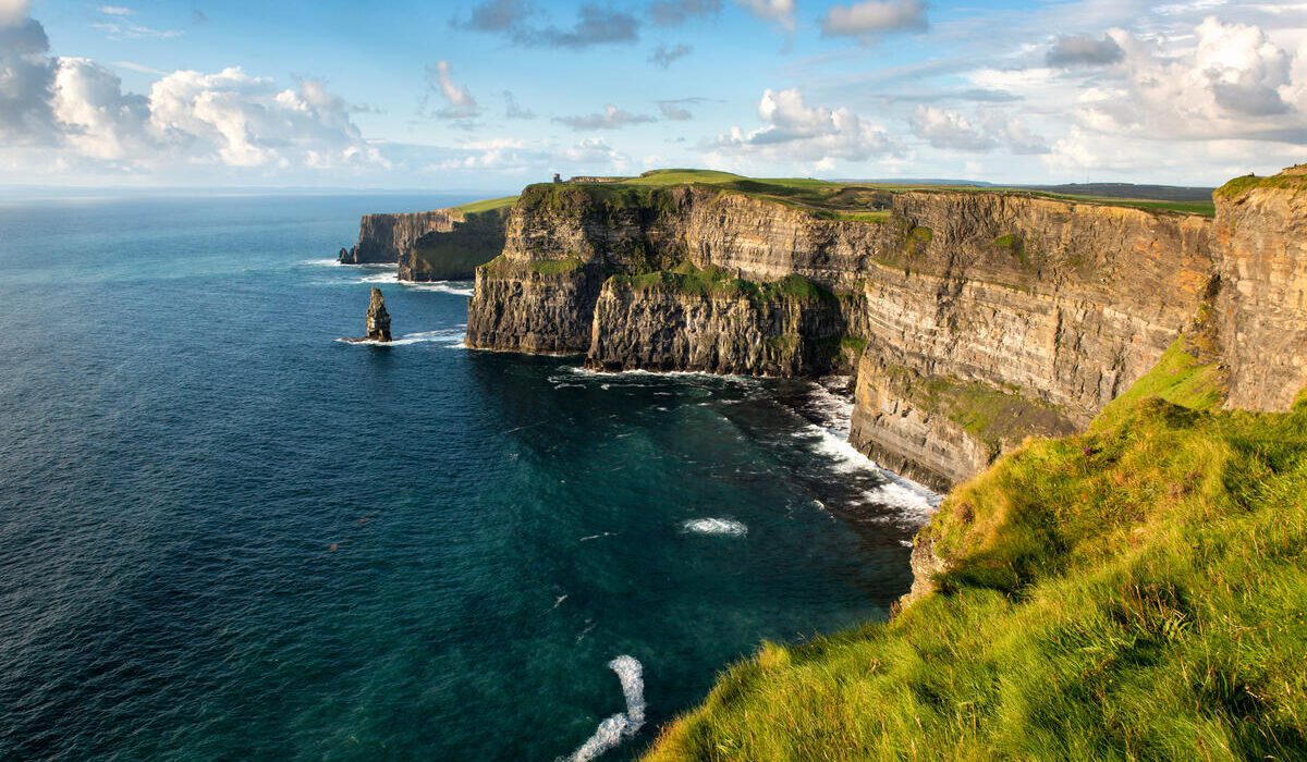Cliffs rise steeply above the Atlantic Ocean, with green grass on top and waves crashing below under a partly cloudy sky.