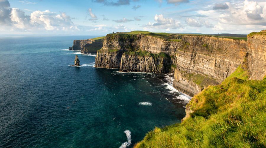 Cliffs rise steeply above the Atlantic Ocean, with green grass on top and waves crashing below under a partly cloudy sky.