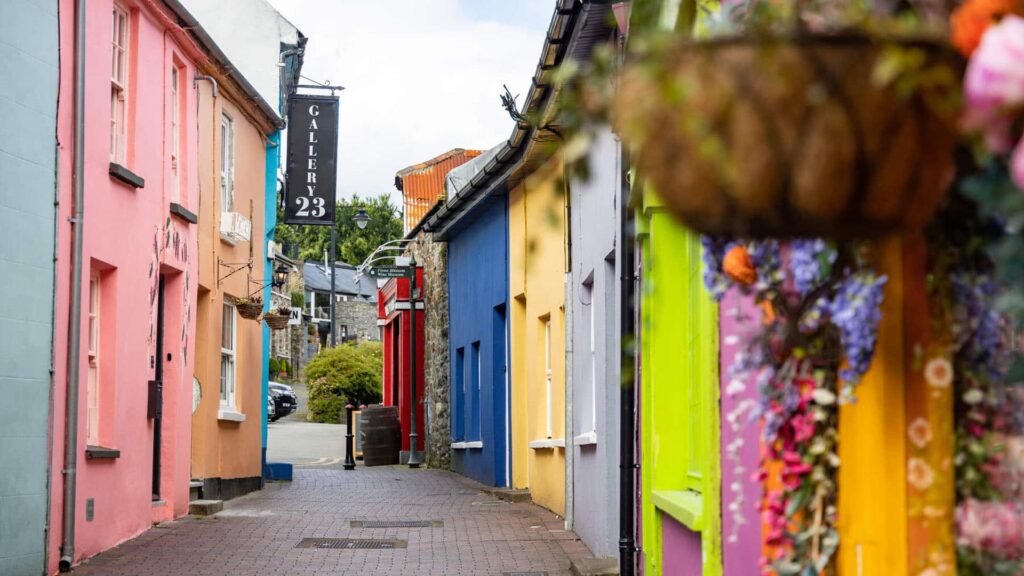 A narrow cobblestone street lined with colorful buildings, including pink, blue, and yellow facades; a sign reads “Gallery 29.” Flowers hang in a basket on the right.