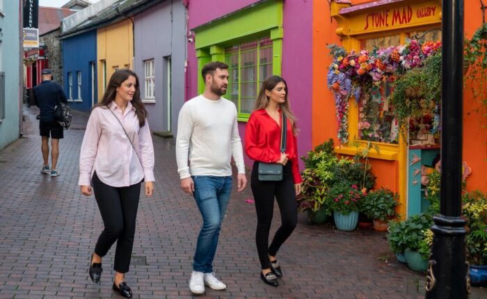 Three people walk past brightly painted shops and a flower-adorned storefront on a cobblestone street.