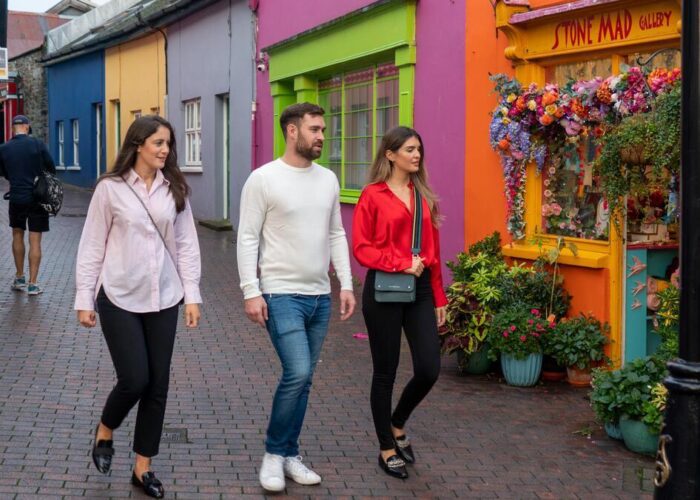 Three people walk past brightly painted shops and a flower-adorned storefront on a cobblestone street.