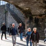 A group of people walk under a stone archway on a sunny day, with stone walls and a metal staircase in the background.