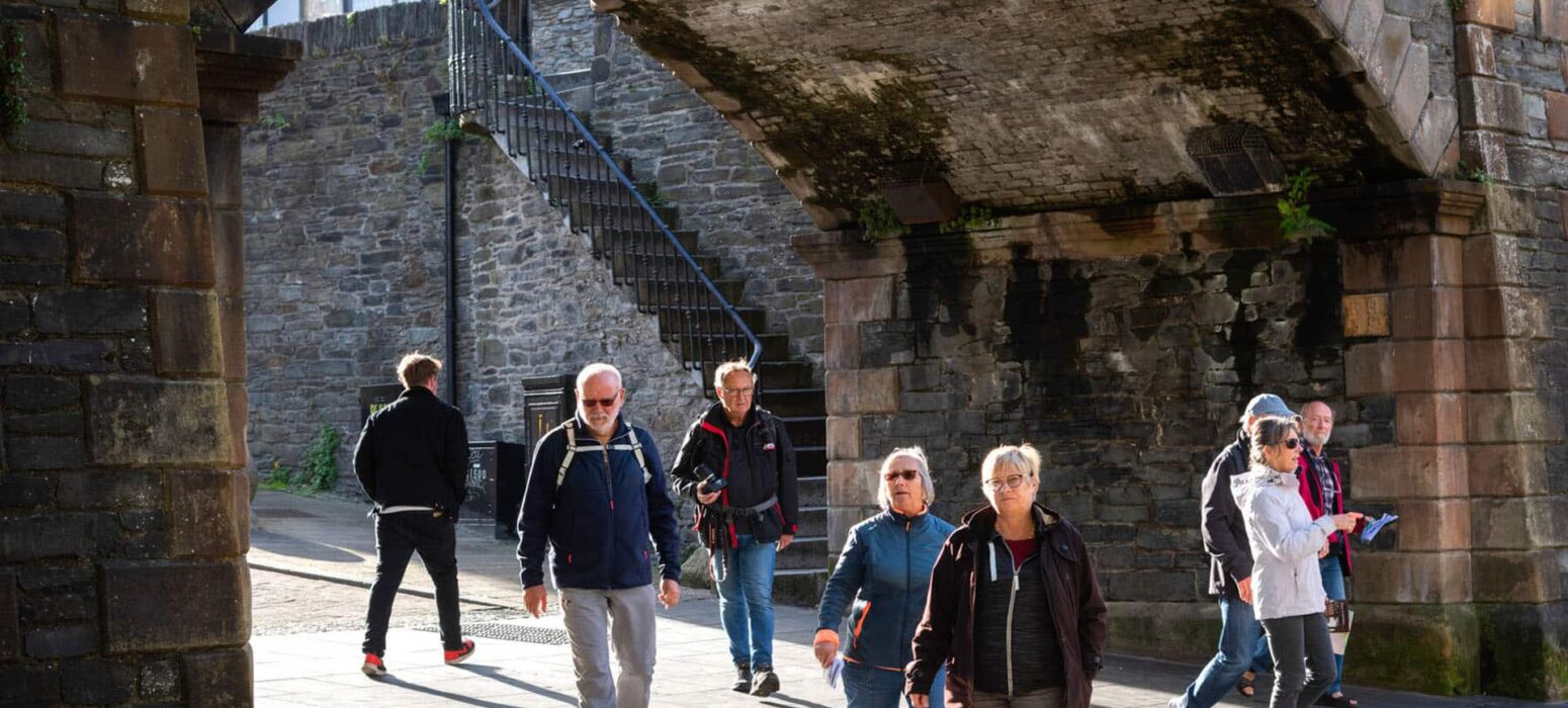 A group of people walk under a stone archway on a sunny day, with stone walls and a metal staircase in the background.