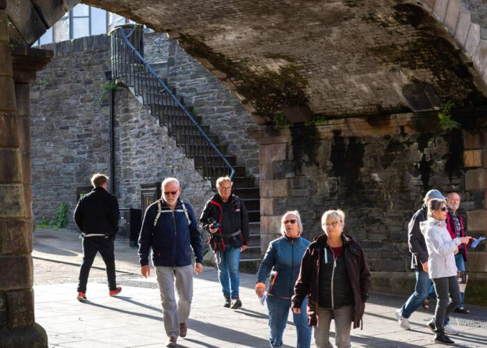 A group of people walk under a stone archway on a sunny day, with stone walls and a metal staircase in the background.