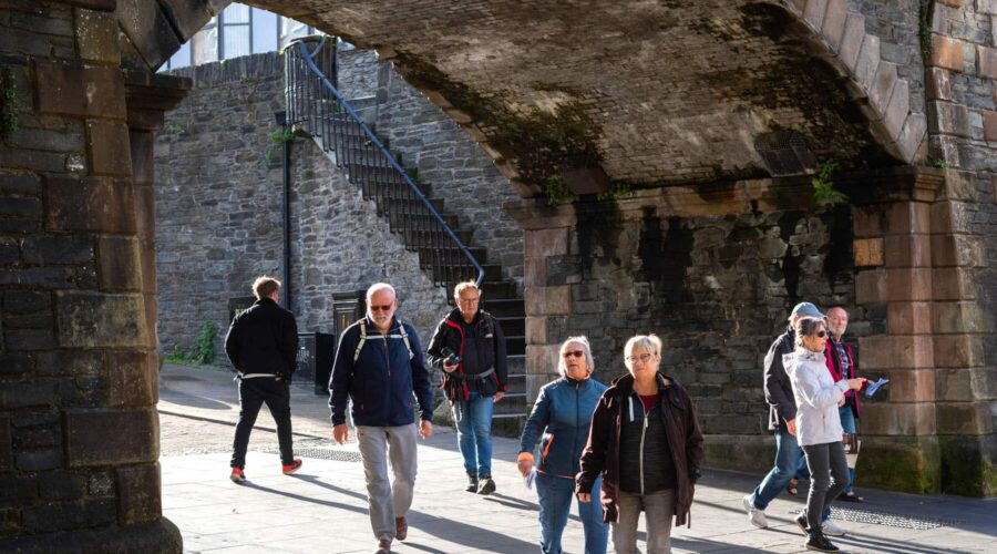 A group of people walk under a stone archway on a sunny day, with stone walls and a metal staircase in the background.