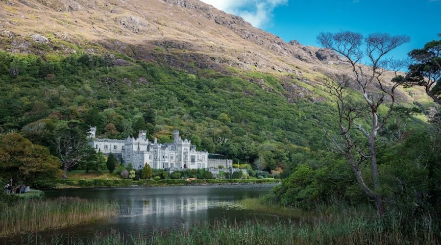 A large grey stone castle with multiple turrets sits at the base of a green hillside, reflected in a calm lake surrounded by trees—an iconic sight on Ireland West tours or South Ireland tours.