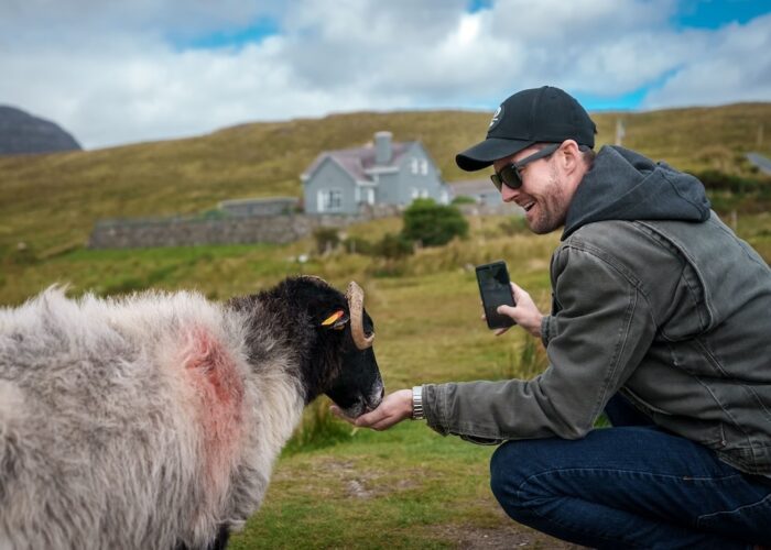 A man in sunglasses kneels and feeds a sheep in a grassy field with a house and hills behind—capturing the charm often found on Ireland West tours—while holding his smartphone.