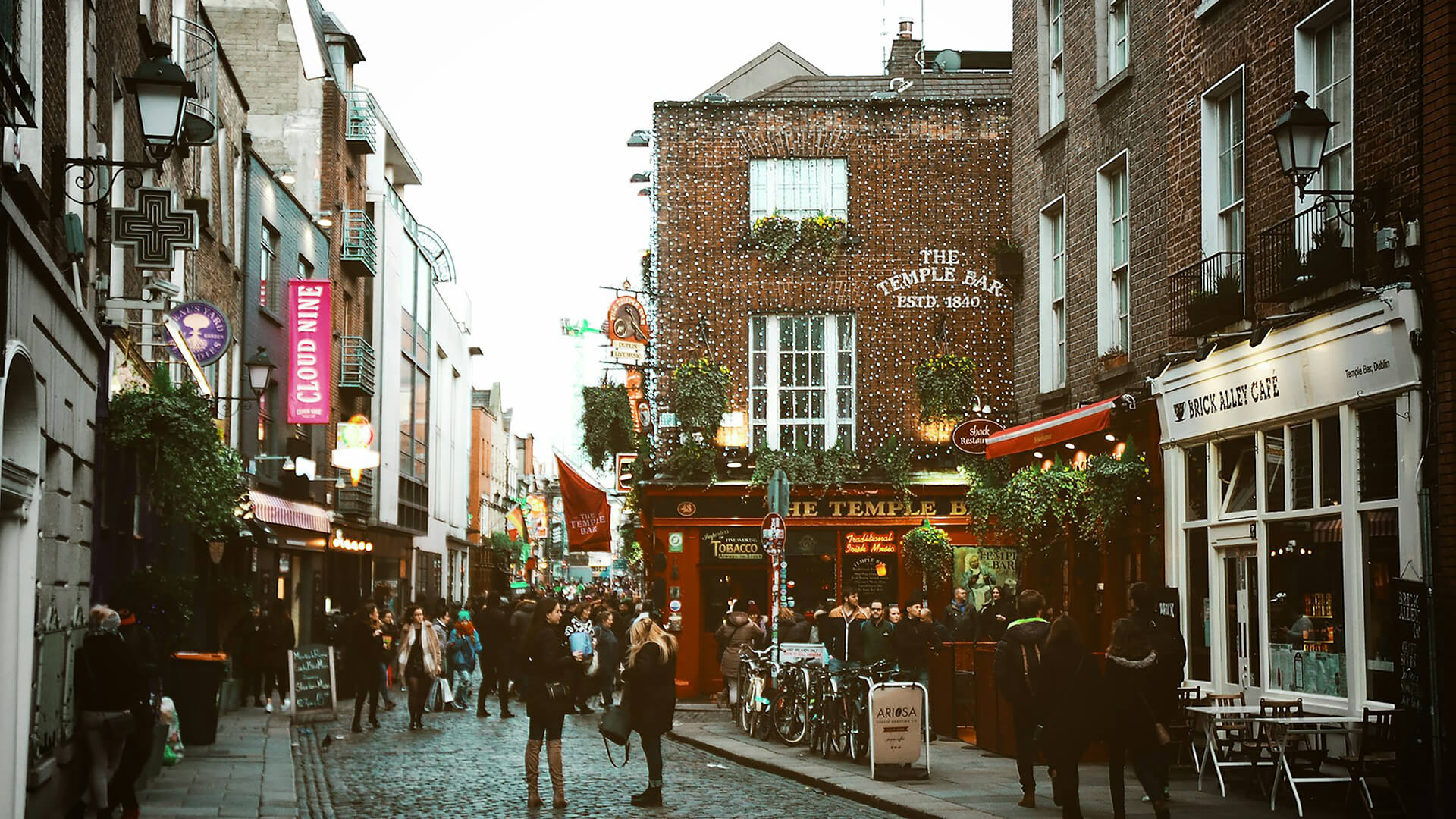 A busy cobblestone street with people walking past shops and cafes, featuring The Temple Bar pub with bikes parked outside and string lights on the building—a classic scene for those enjoying day tours from Dublin Ireland.