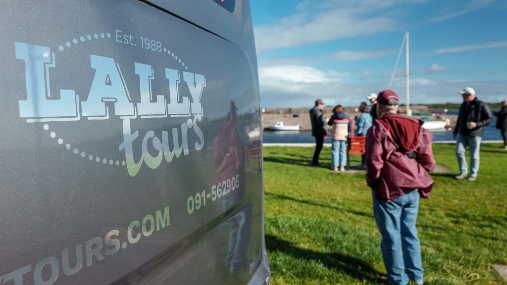 A group of people stands near a body of water and boats, beside a van displaying the "Lally Tours" logo and contact information.