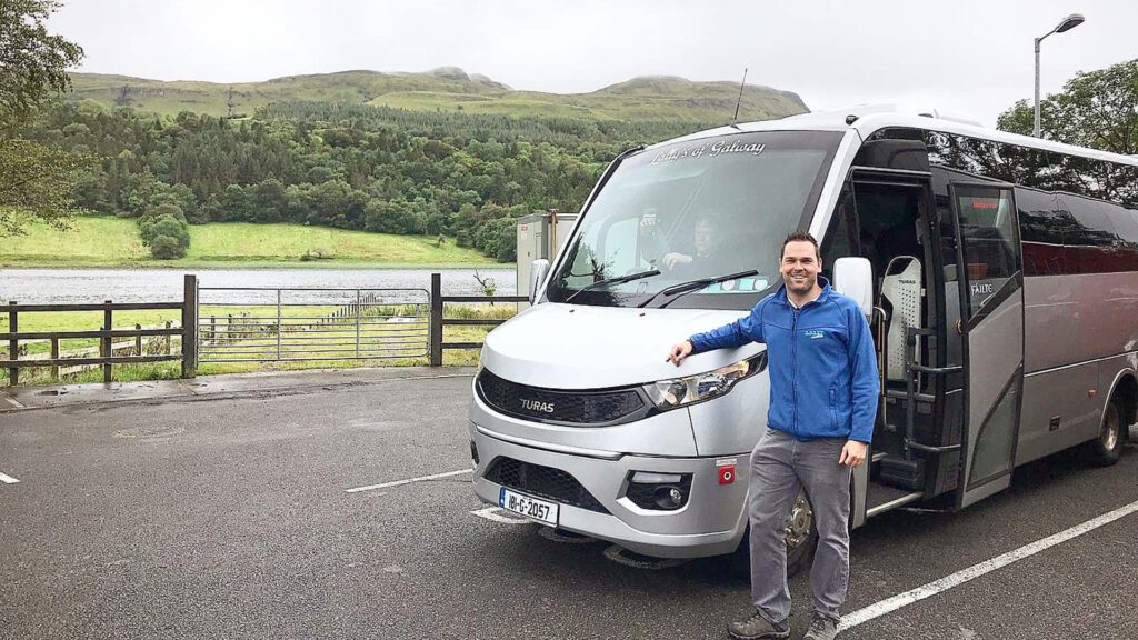 A man in a blue jacket stands beside a parked silver tour bus in a lot with trees and hills in the background.