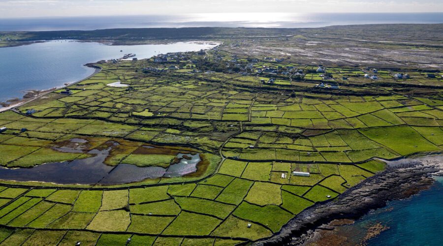 Aerial view of a coastal landscape near Galway with patchwork green fields, stone fences, scattered houses, and the sea in the background under a partly cloudy sky—perfect for a Guided Day Tour to Inis Mór.