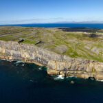 Aerial view of rocky cliffs and green fields meeting the ocean on Inis Mór, with calm blue water and clear sky in the background—discover this stunning landscape on a guided day tour from Galway.