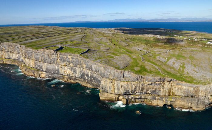 Aerial view of rocky cliffs and green fields meeting the ocean on Inis Mór, with calm blue water and clear sky in the background—discover this stunning landscape on a guided day tour from Galway.