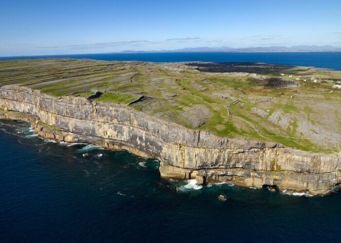 Aerial view of rocky cliffs and green fields meeting the ocean on Inis Mór, with calm blue water and clear sky in the background—discover this stunning landscape on a guided day tour from Galway.