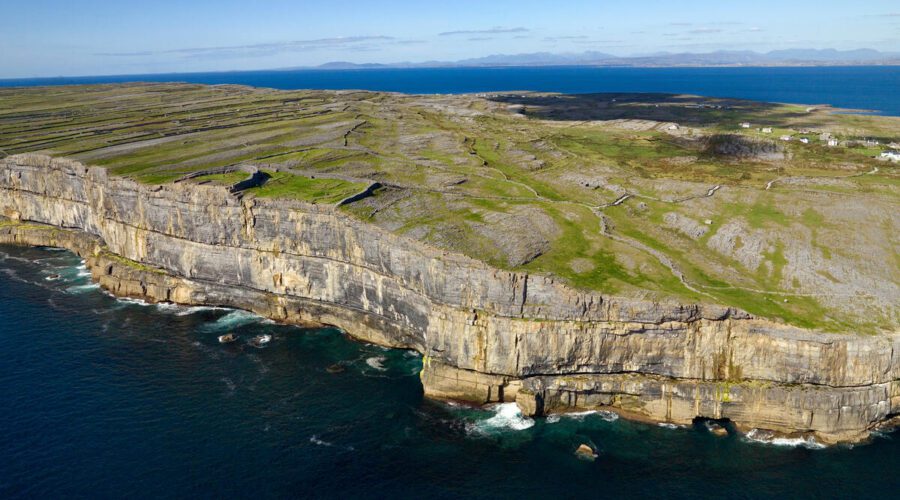 Aerial view of rocky cliffs and green fields meeting the ocean on Inis Mór, with calm blue water and clear sky in the background—discover this stunning landscape on a guided day tour from Galway.
