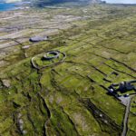 Aerial view of Inis Mór’s green, stone-walled landscape with two ancient stone forts and a distant coastline under a blue sky—discover its beauty on a Guided Day Tour from Galway.