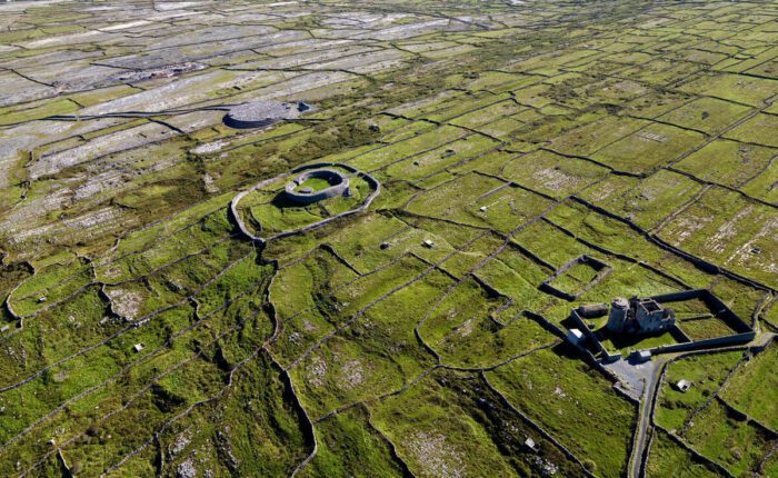 Aerial view of Inis Mór’s green, stone-walled landscape with two ancient stone forts and a distant coastline under a blue sky—discover its beauty on a Guided Day Tour from Galway.